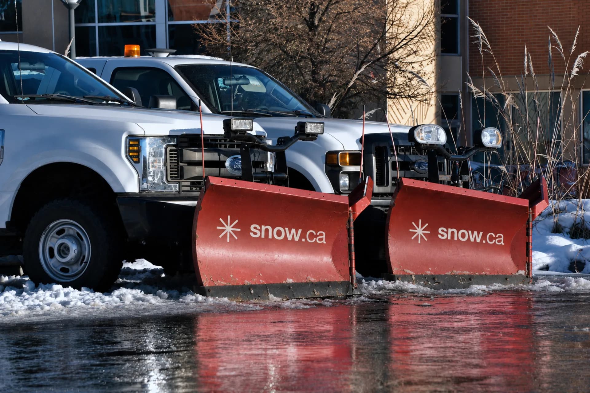 Two snow.ca Ford F-Series plow trucks with branded red plow blades parked at a Canadian commercial site after a winter storm — snow.ca national snow management fleet.