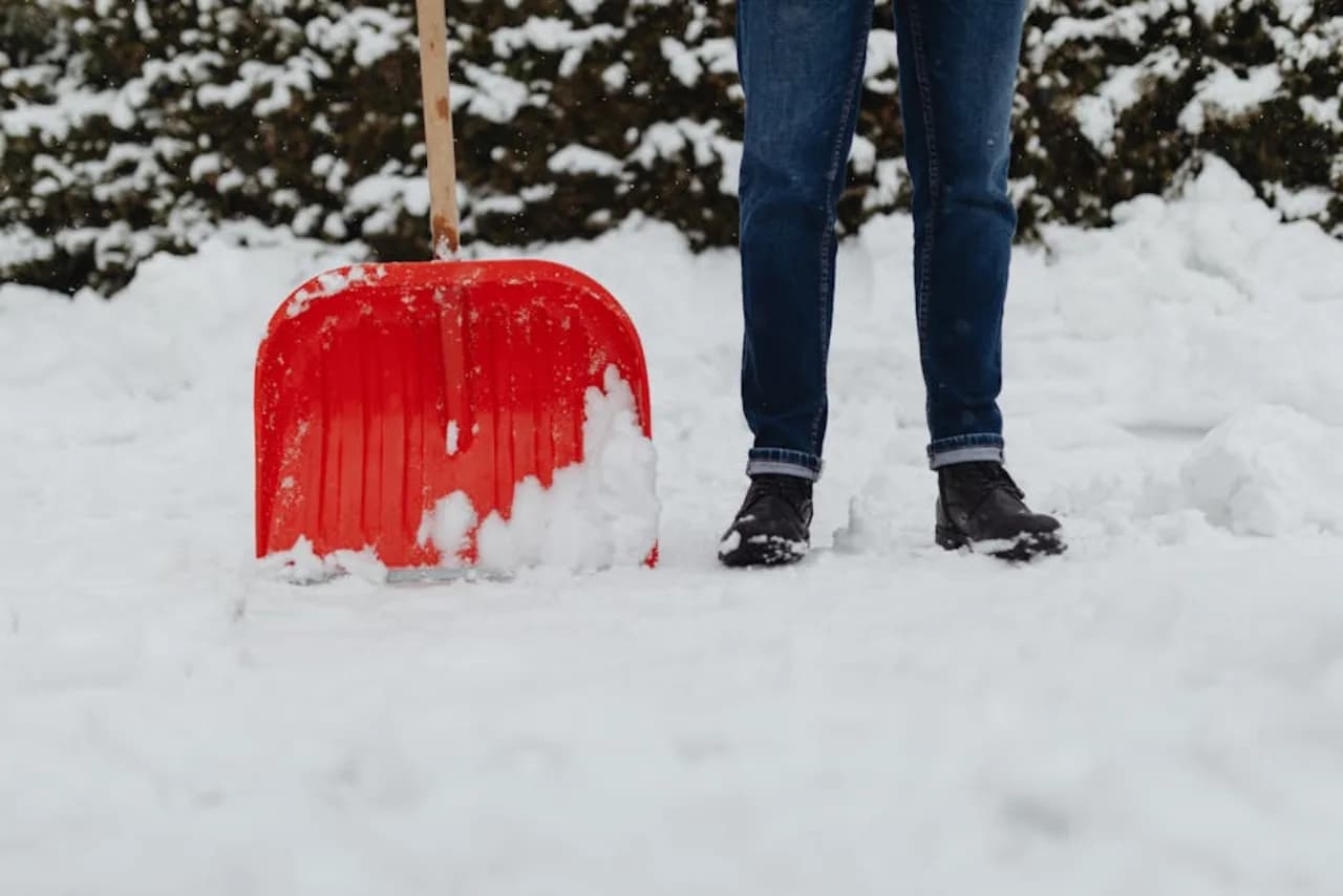 Une pelle à neige rouge appuyée sur une entrée enneigée canadienne avec l’opérateur à côté — contexte procédural pour les guides pratiques snow.ca destinés aux propriétaires.